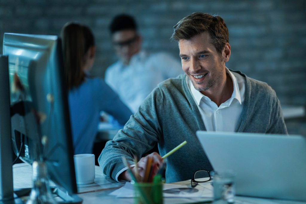 happy-male-entrepreneur-using-desktop-pc-while-working-office-there-are-people-background